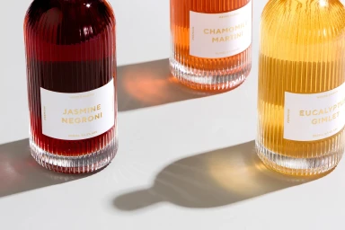 Three fluted glass bottles with white labels containing different coloured craft cocktails on a white surface.