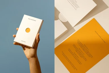 Hand holding a white product box with a circular cutout against a blue background, next to printed paper materials