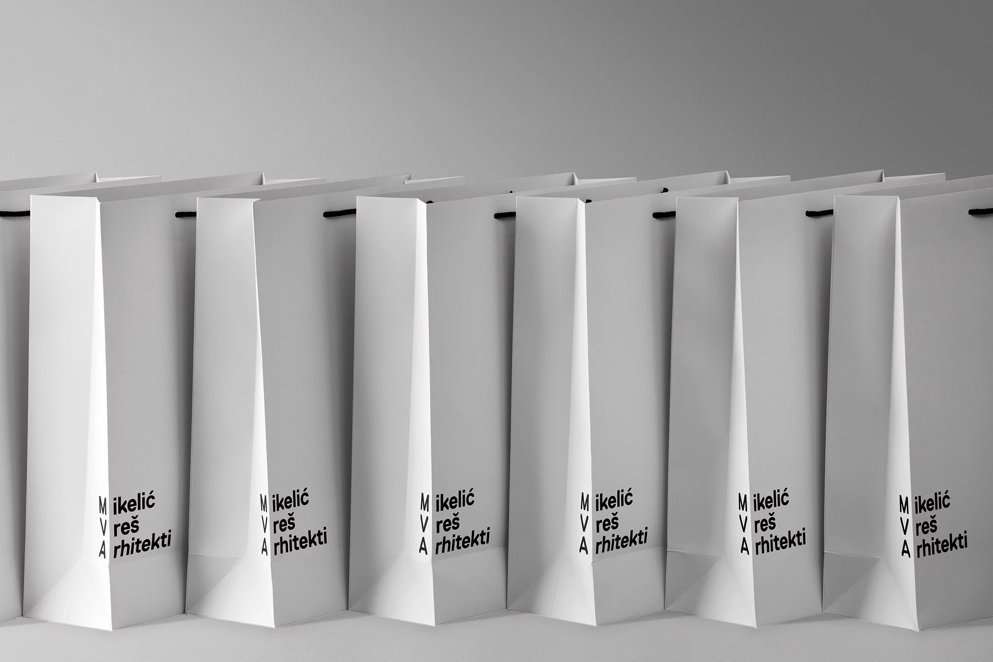 Row of identical white shopping bags with black handles, arranged in a repeating pattern against a gradient gray background, studio lighting.