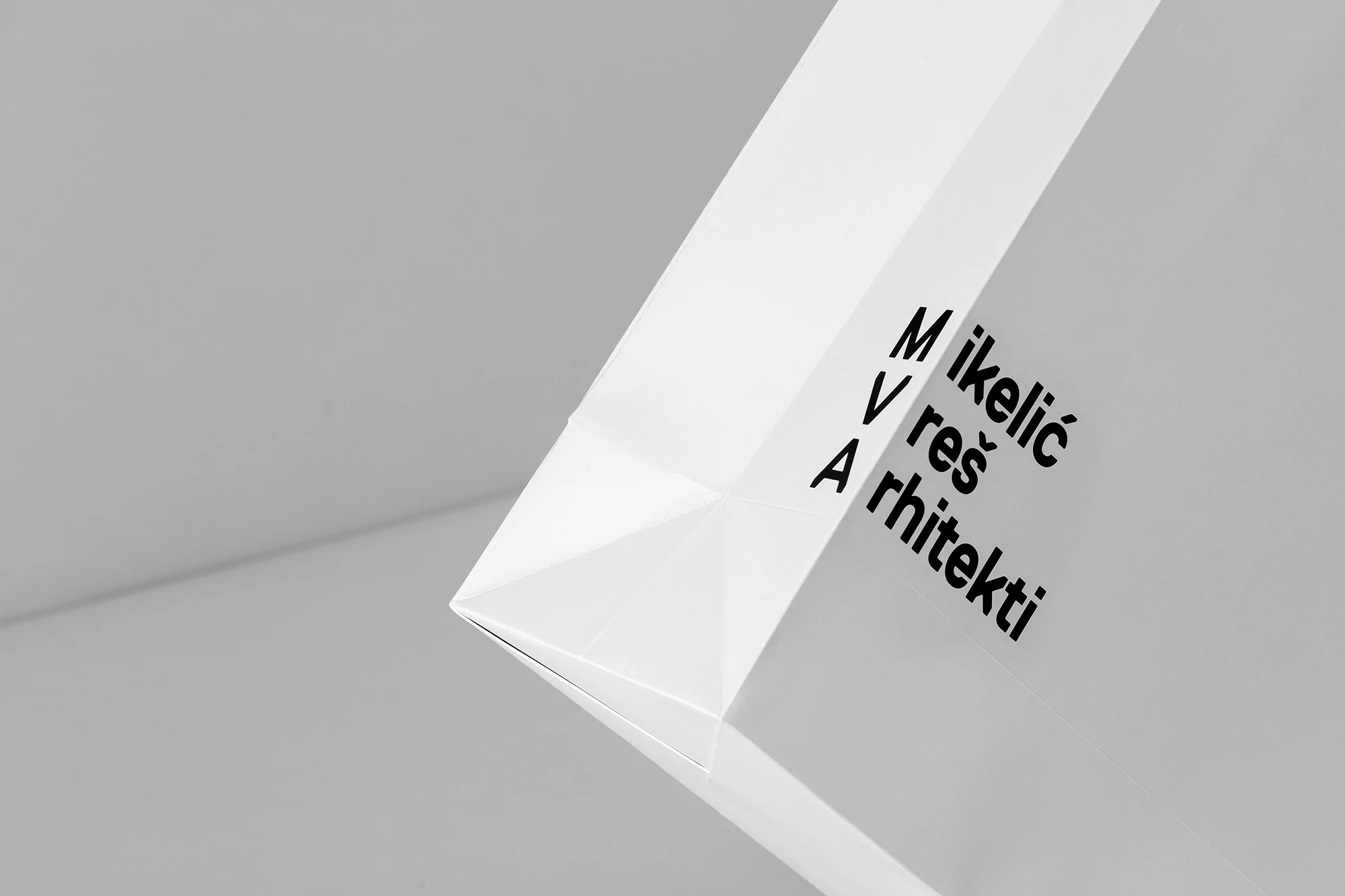 Minimalist close-up of white paper bag with black sans-serif text on a gray surface, emphasizing graphic design and architectural branding.