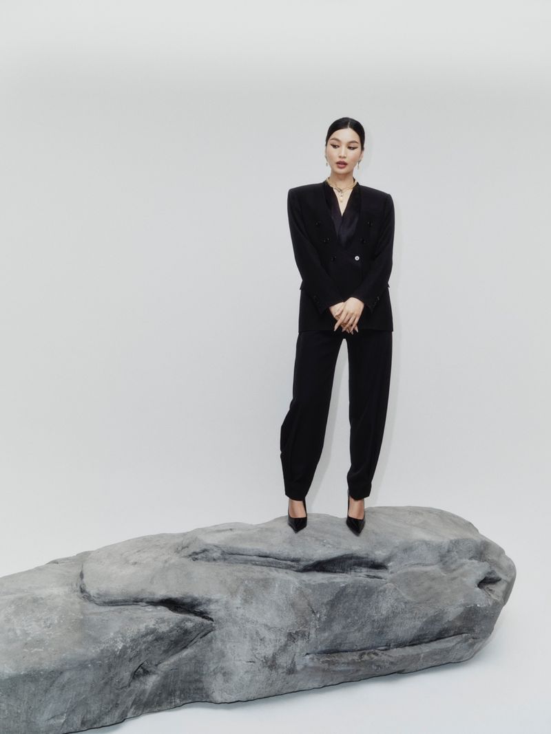 Elegant Asian woman in a black suit and high heels poses atop a gray rock in a bright studio setting, creating a striking portrait.