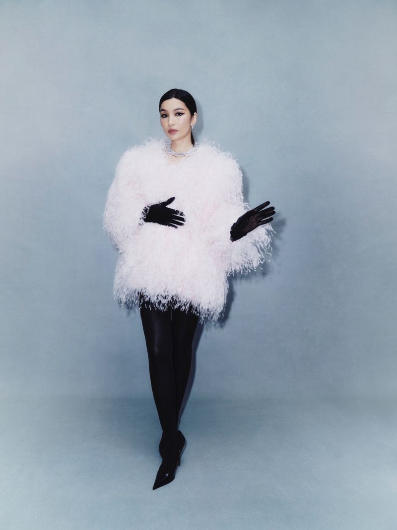 Adult woman poses in feathered coat, black gloves, tights, and heels against a plain background in a fashion-style photograph.
