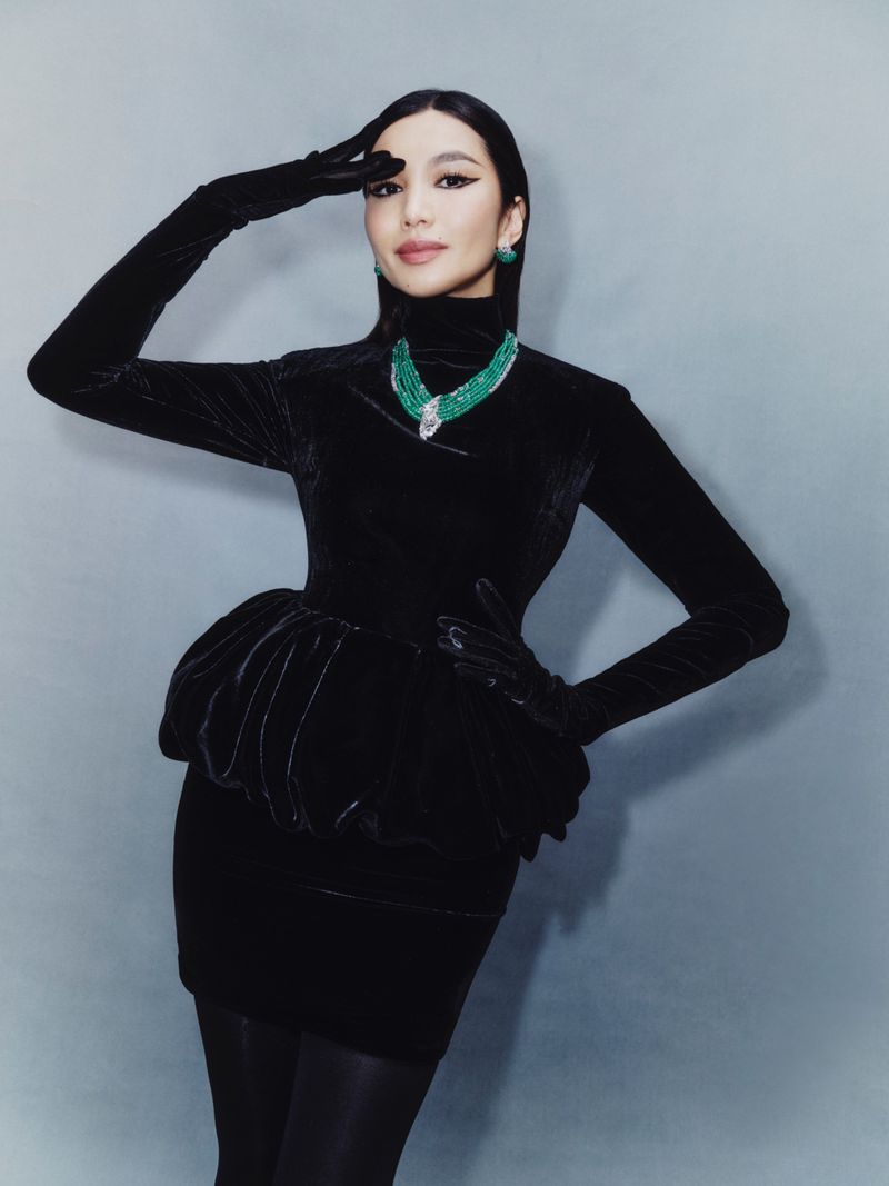 Elegant woman in black velvet dress, long gloves, and emerald jewelry strikes a pose against a soft grey backdrop in a fashion portrait.