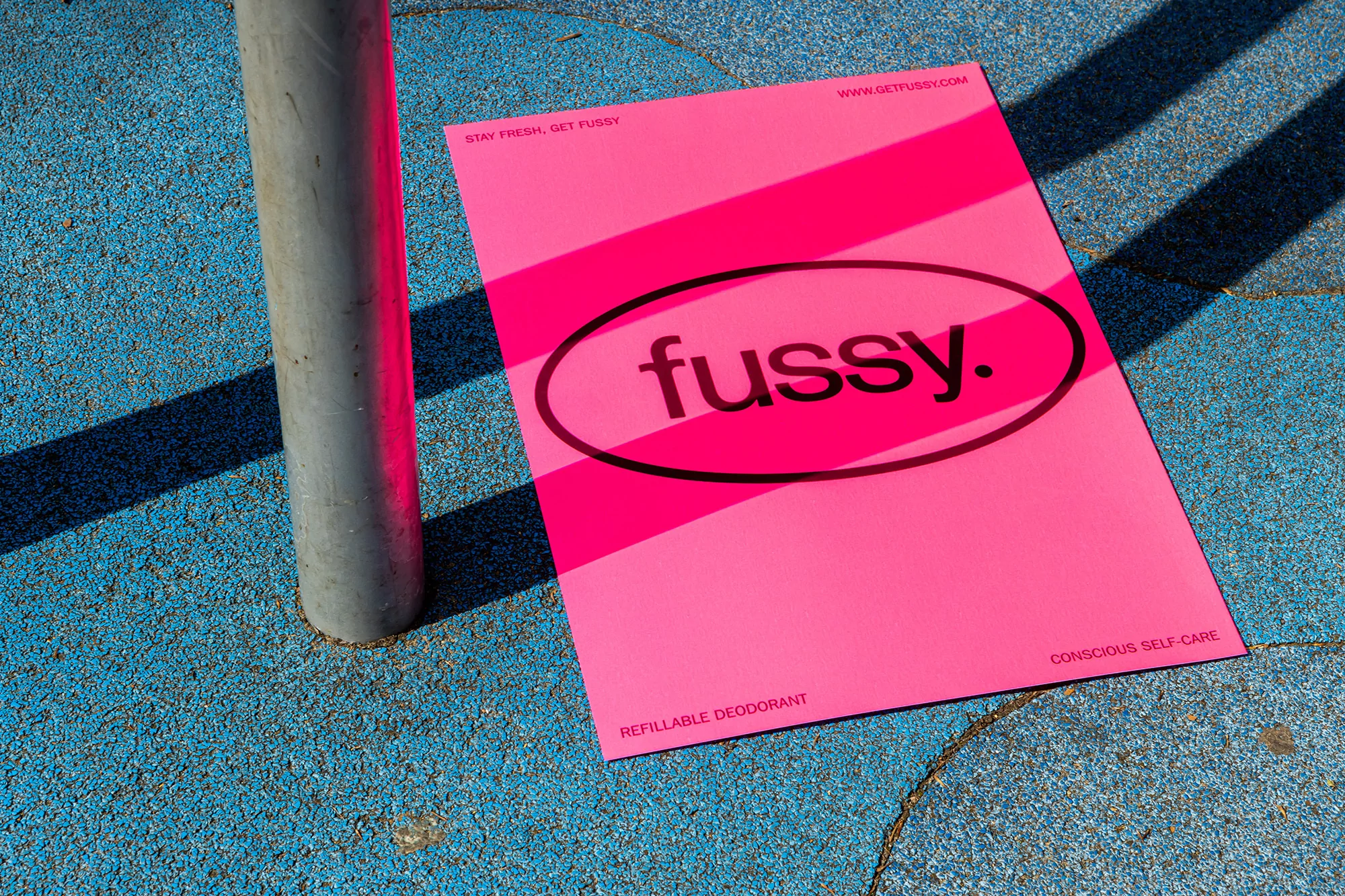 Bright pink poster on a textured blue surface featuring the black logo 'fussy.' and taglines, 'Stay Fresh, Get Fussy' and 'Conscious Self-Care'.