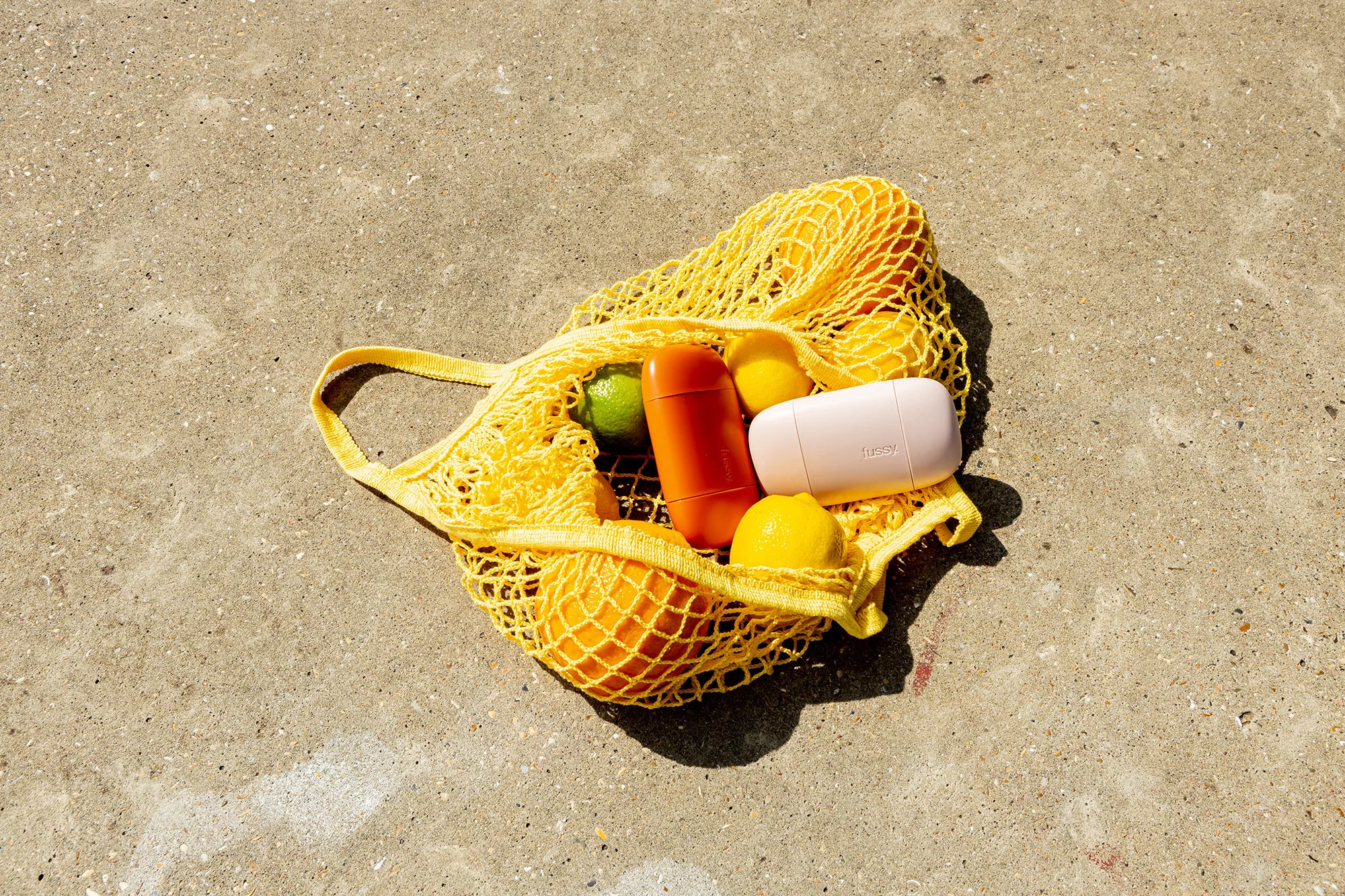 Overhead shot of a yellow string bag filled with oranges, lemons, and personal care containers on a concrete surface outdoors.