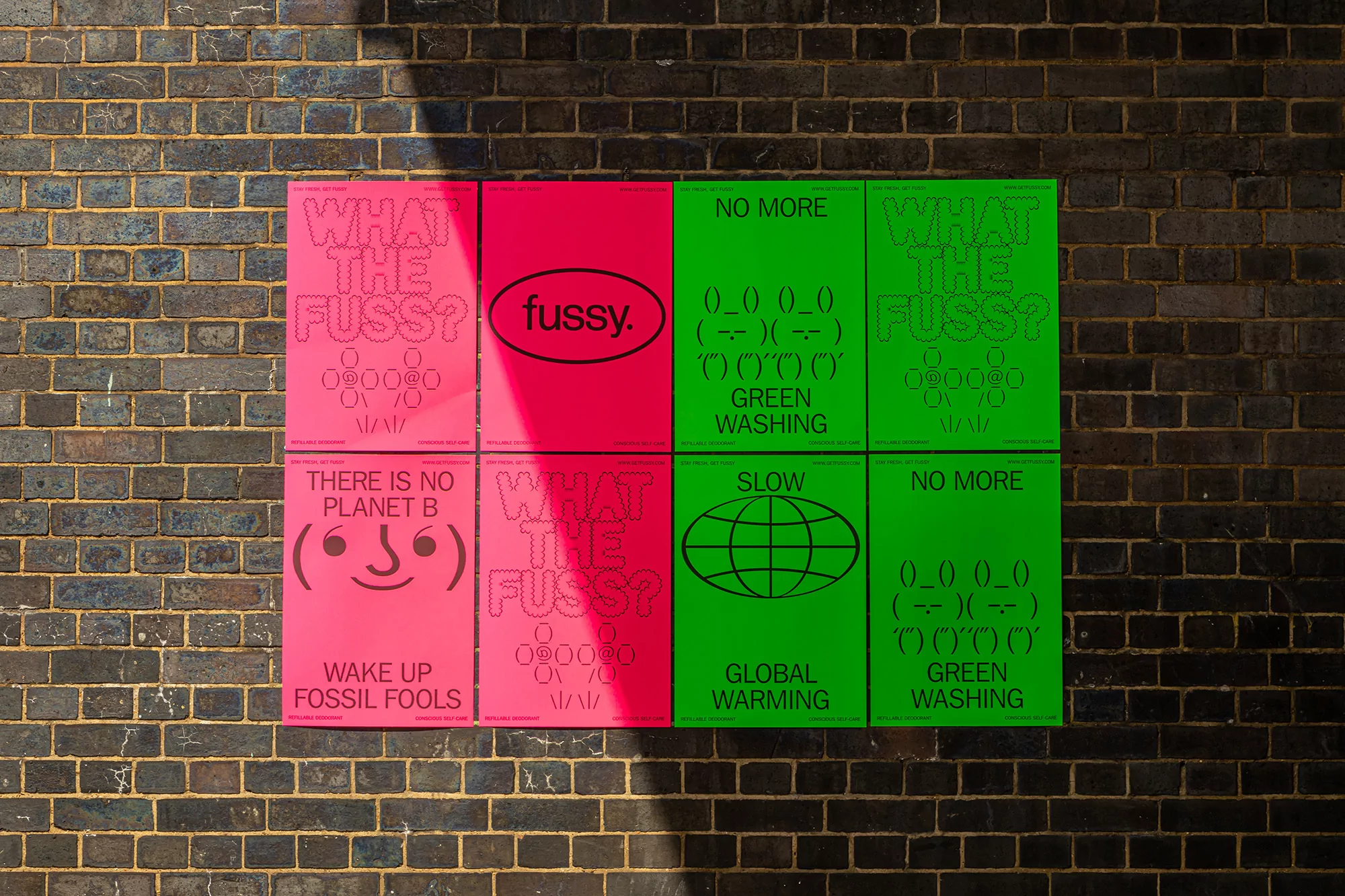 Eight environmental awareness posters in pink and green with text and illustrations are fixed to a weathered brick wall in sunlight.