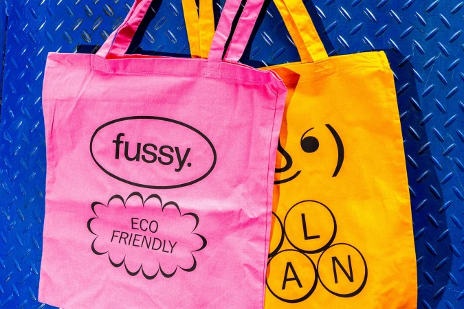 Pink and yellow tote bags with eco-friendly designs sit on a blue metal background, ideal for sustainable living and reusable shopping.