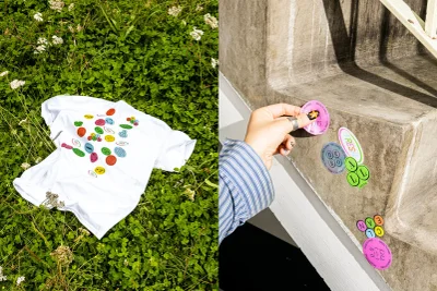 White t-shirt with colourful stickers lying on green clover, and a hand placing a sticker on a concrete wall
