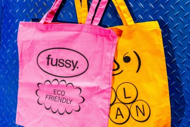 Bright pink and yellow canvas tote bags featuring bold black graphic typography against a blue metal background