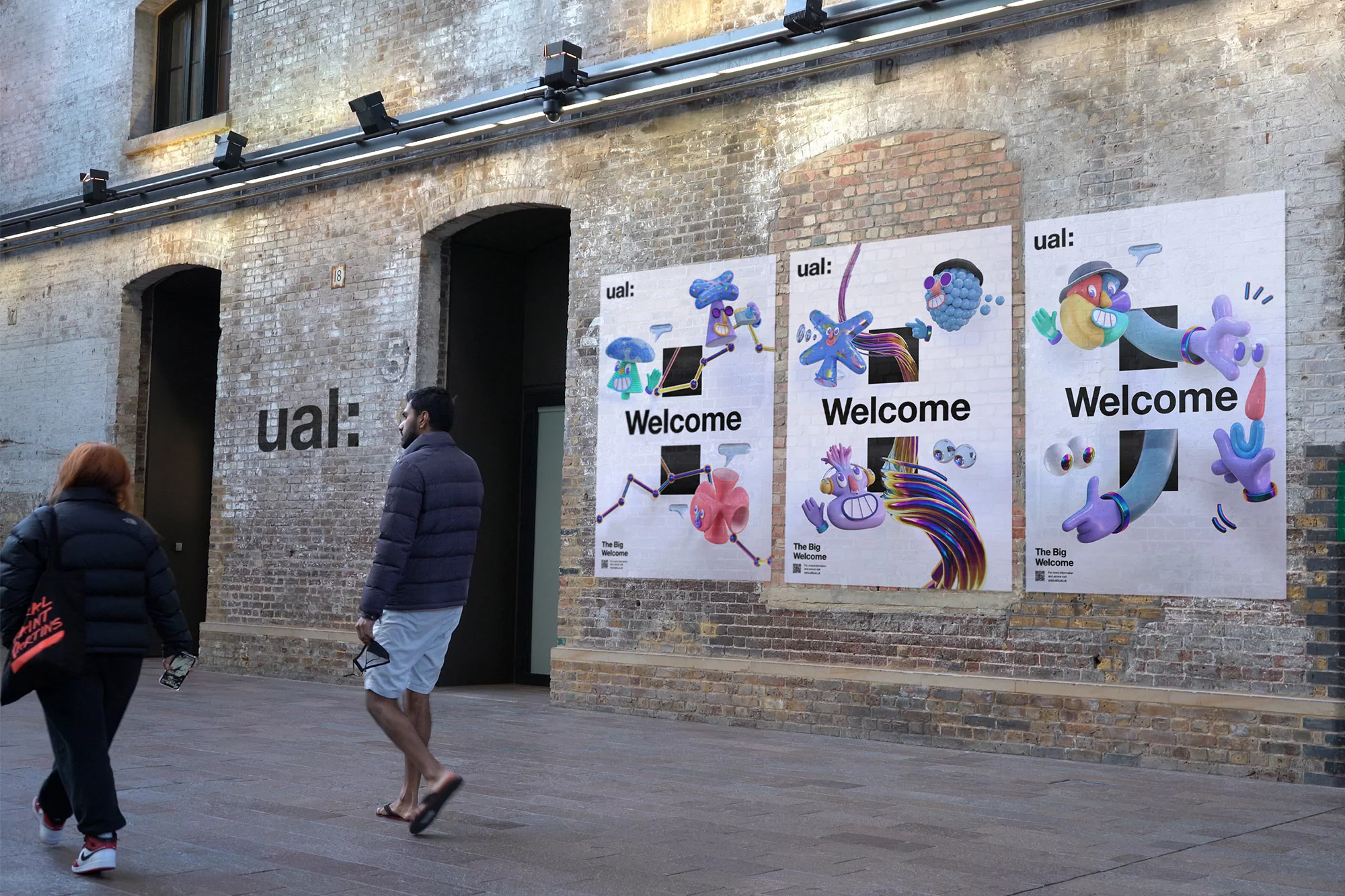 Three posters on a brick wall showcase UAL's Big Welcome campaign with cartoonish figures and the word 'Welcome', viewed from a city sidewalk.