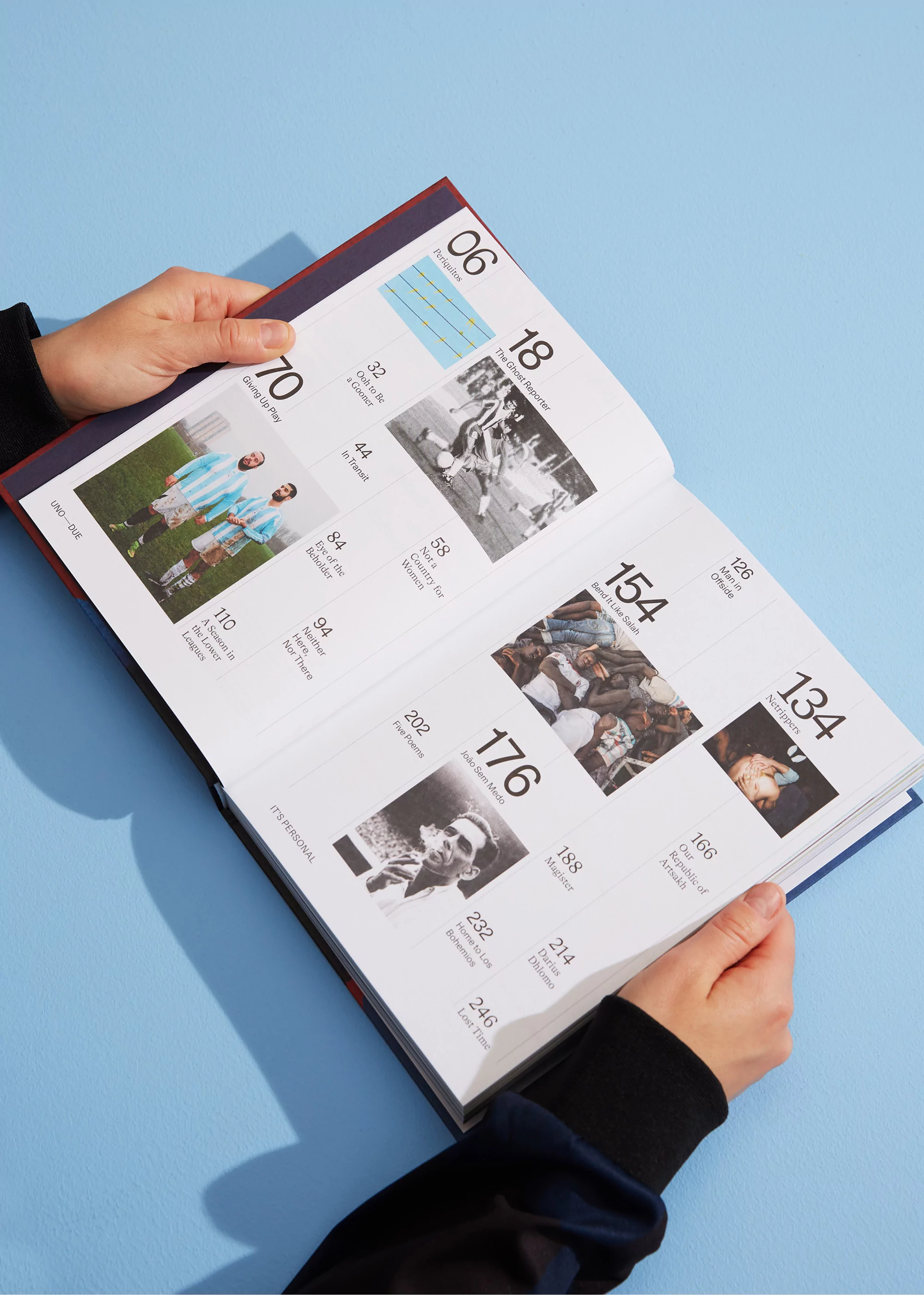 Open book with photographs and page numbers held by two hands, against a blue backdrop. The book displays black and white and color imagery.