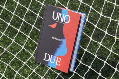 Hardcover book with red, black, and blue cover resting on a white goal net over green artificial turf