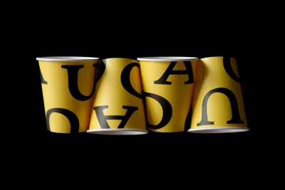 Four yellow paper coffee cups with large black typographic lettering arranged in a row against a solid black background.