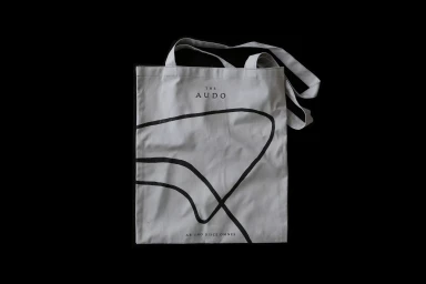 Off-white cotton canvas tote bag with black branding and curved graphic lines displayed against a solid dark background.