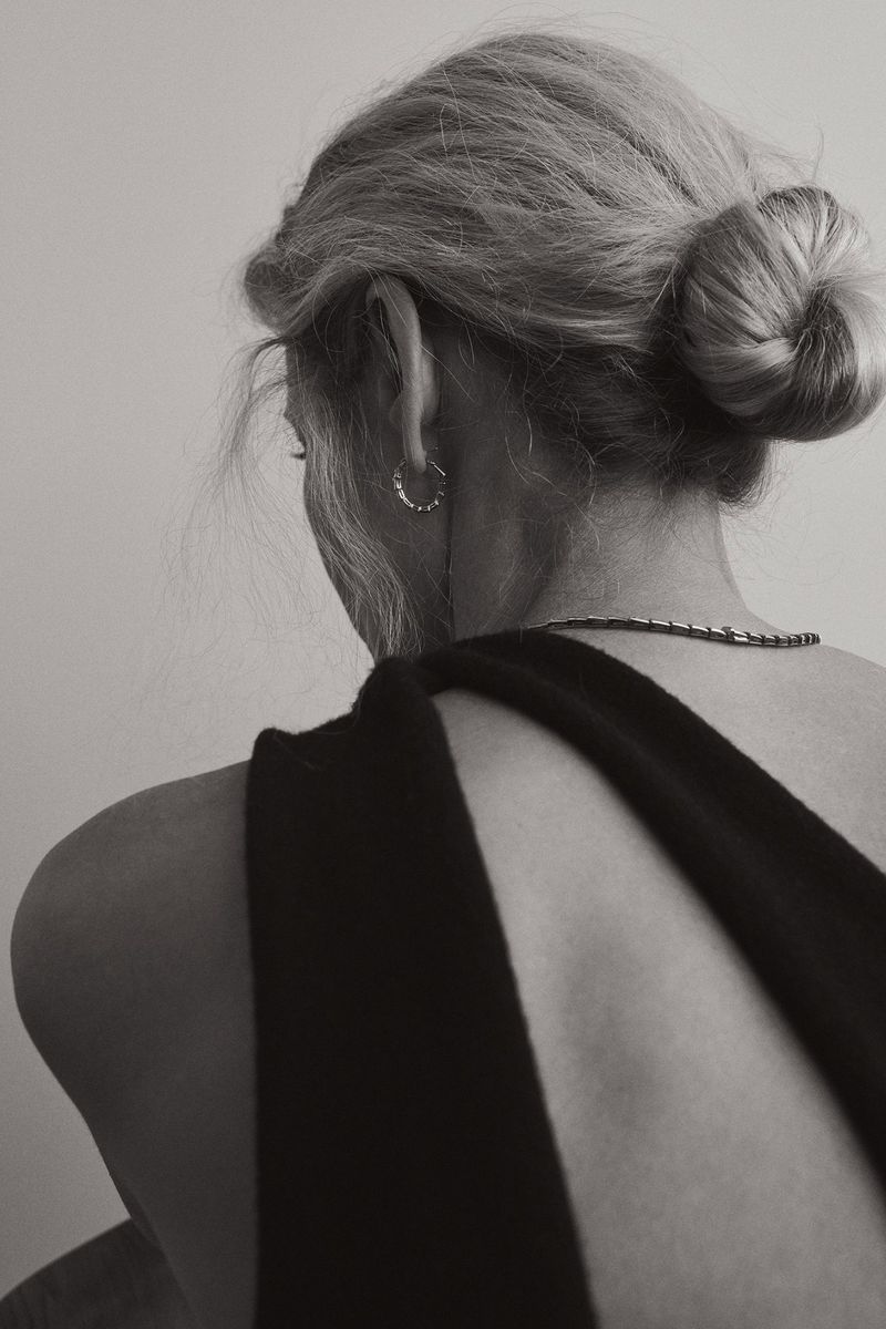 Back view of a woman with blonde hair in a bun, wearing a black dress. Simplistic aesthetic in a softly lit neutral setting.