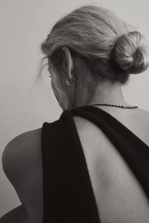Back view of a woman with blonde hair in a bun, wearing a black dress. Simplistic aesthetic in a softly lit neutral setting.