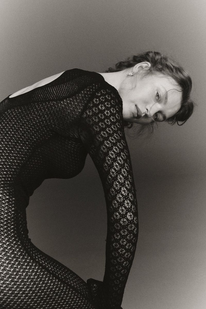 Black and white portrait of a woman in a patterned body suit. She's leaning forward with her head tilted and looking at the camera.