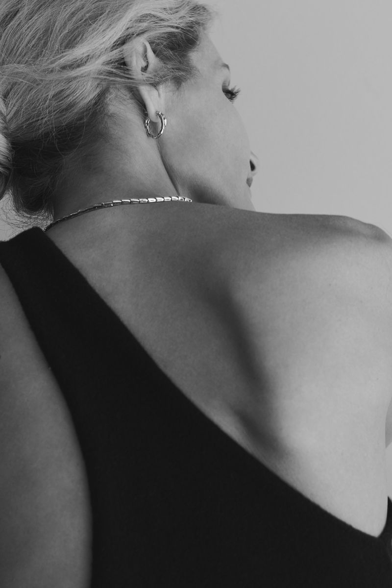 Monochromatic photo of a woman with blonde hair in a low bun, wearing a black one-shoulder top and silver jewelry, showcasing her upper back.