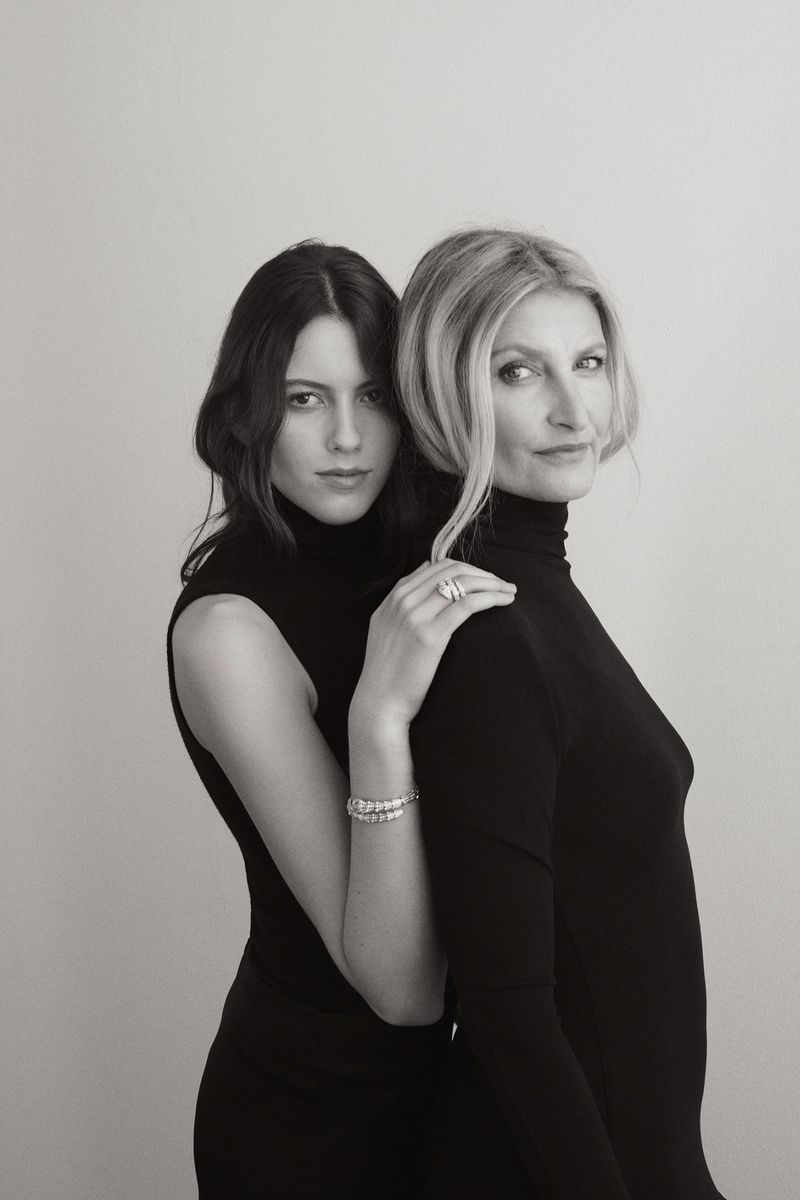 Elegant portrait of a young woman with dark hair and older blond woman in sleek black turtlenecks, posing together against a light background.