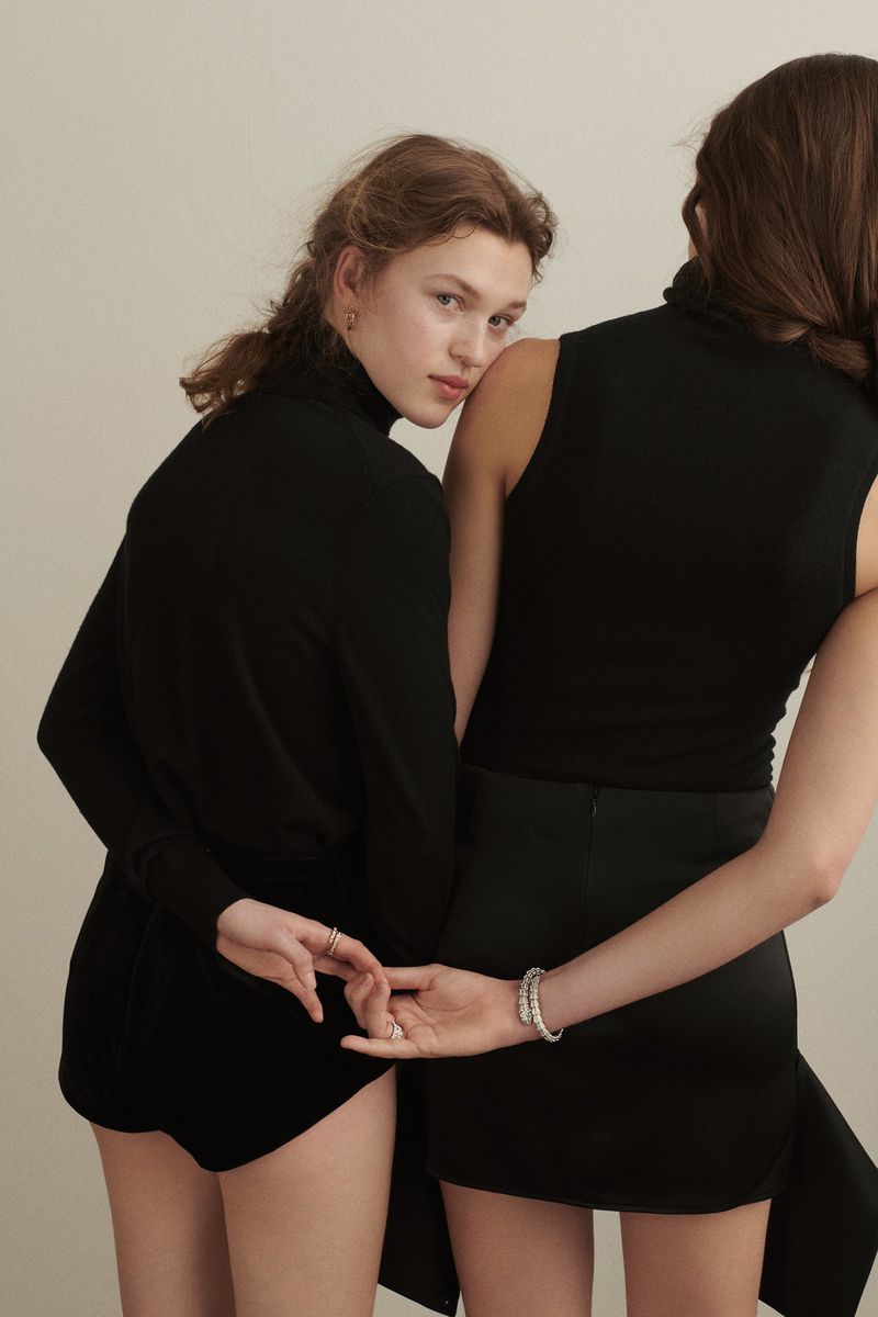 Two women pose back-to-back in black outfits against an off-white backdrop. One woman is facing forward, the other is facing away.