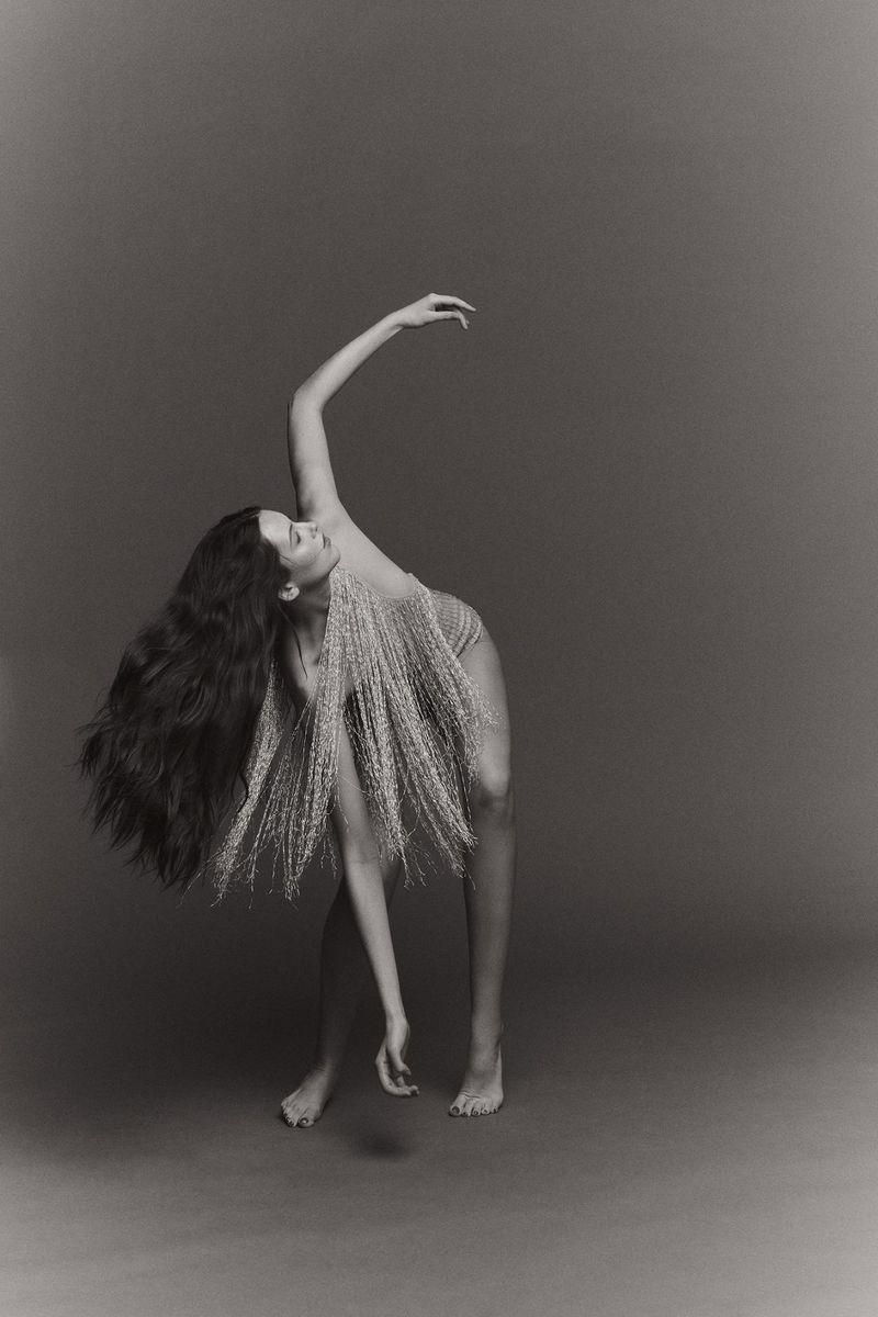 Monochrome photo of a slender adult woman with long wavy hair, adorned in wispy botanical fronds, posing in a curved dance-like posture.