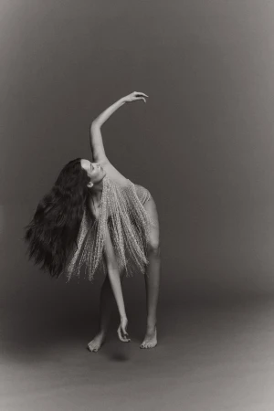 Monochrome photo of a slender adult woman with long wavy hair, adorned in wispy botanical fronds, posing in a curved dance-like posture.