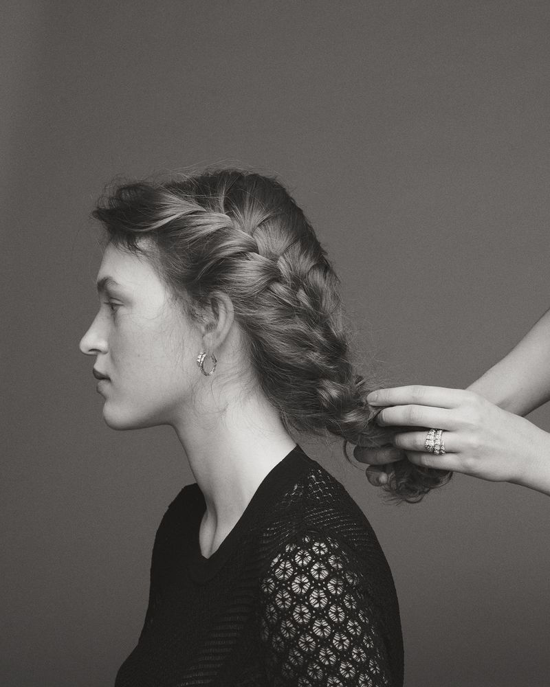 Profile of a woman with a braided hairstyle, being styled by another set of hands in a black and white photograph.