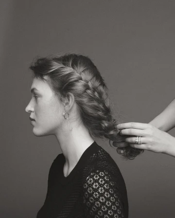 Profile of a woman with a braided hairstyle, being styled by another set of hands in a black and white photograph.