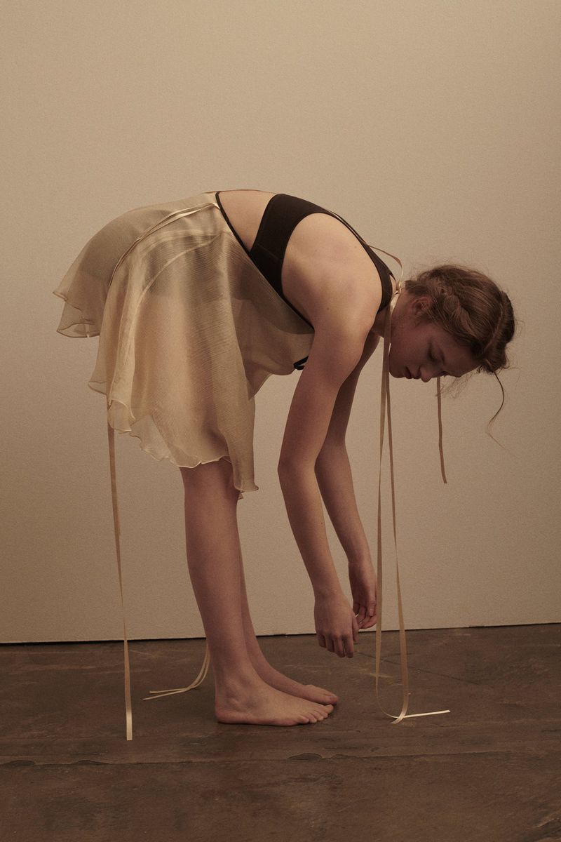 Young woman bends over in a dance pose wearing a beige dress with ribbons in a minimalist studio. Her face is downcast. 