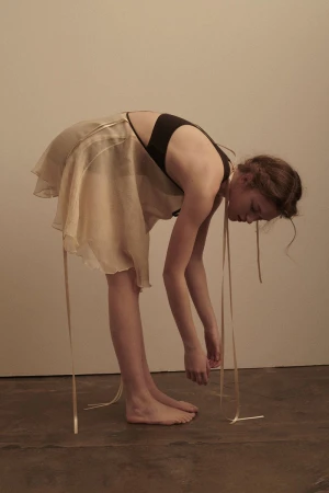 Young woman bends over in a dance pose wearing a beige dress with ribbons in a minimalist studio. Her face is downcast. 