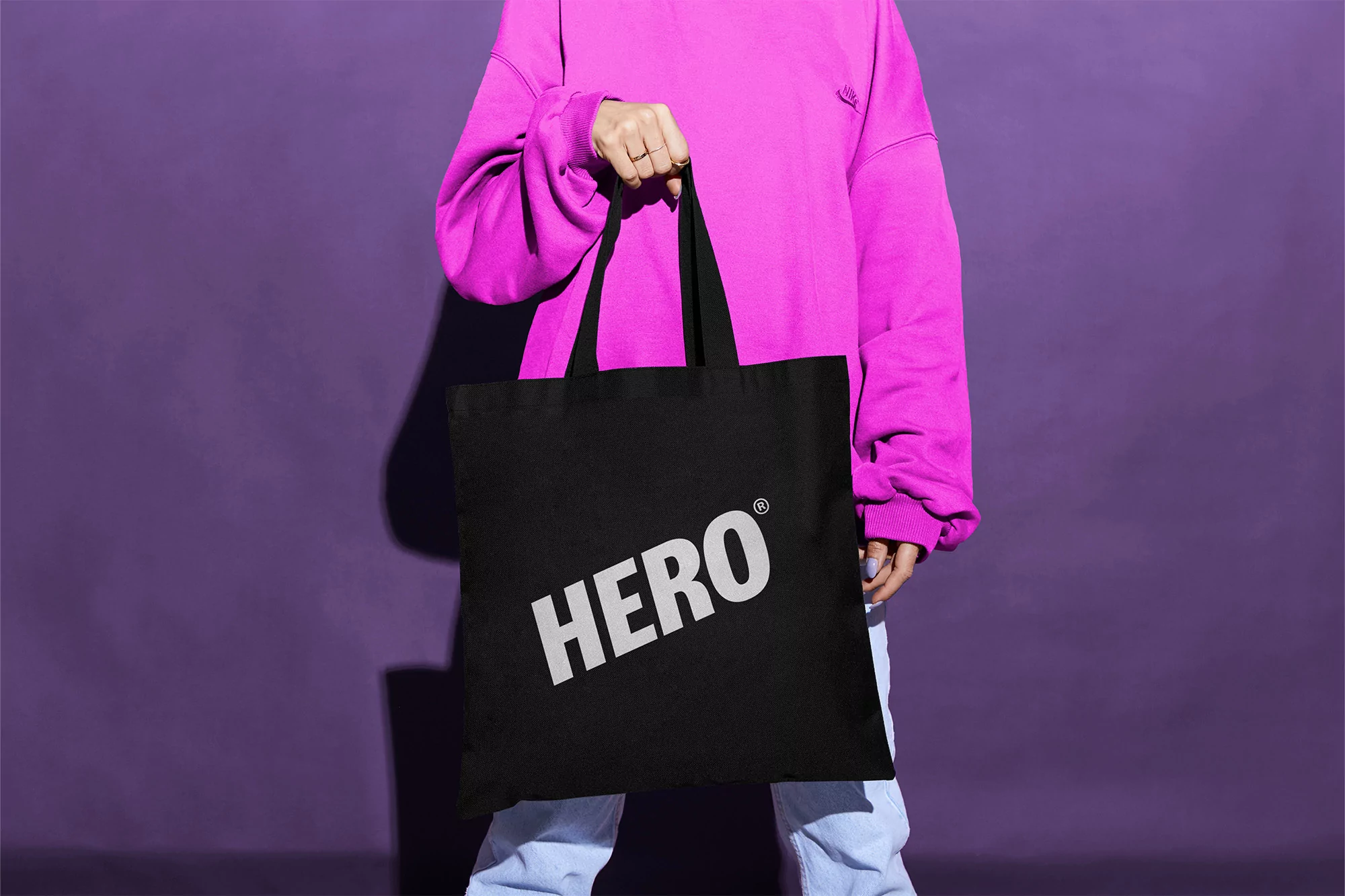 Person holding a black tote bag with the word HERO printed on it, wearing a pink sweater and jeans against a purple background.