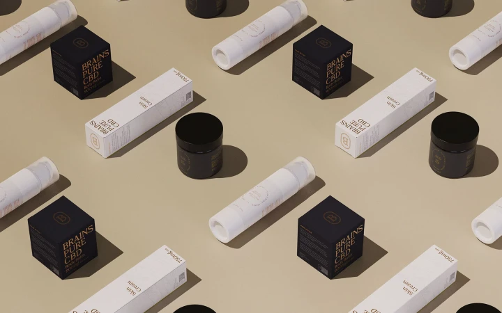 A flatlay arrangement of Brains Pure CBD product packaging including black jars, white boxes, and tubes on a beige surface.