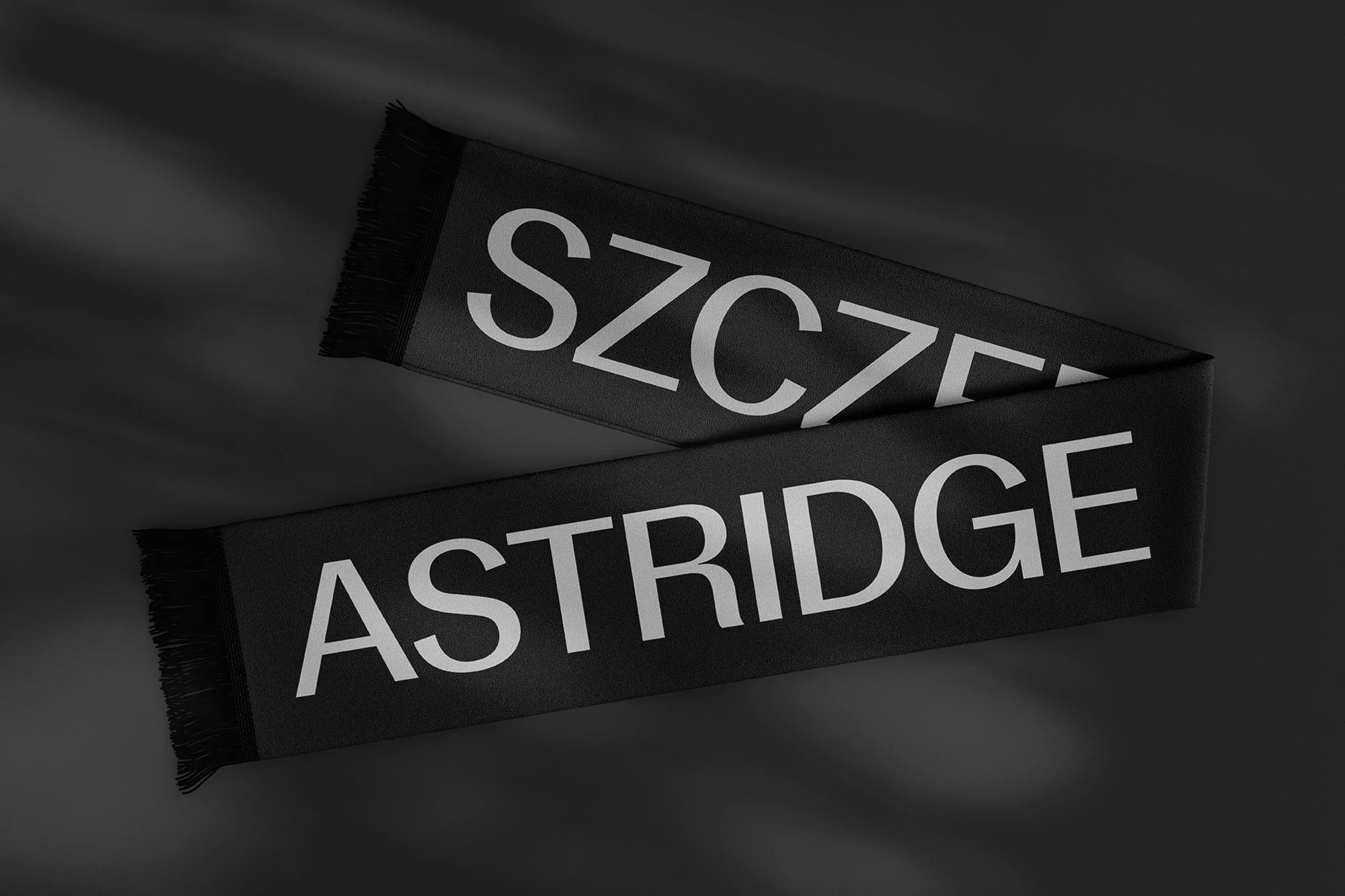 Black scarf with 'Szczelige' and 'Astridge' in white text on a dark surface, featuring fringed edges and shadow play.