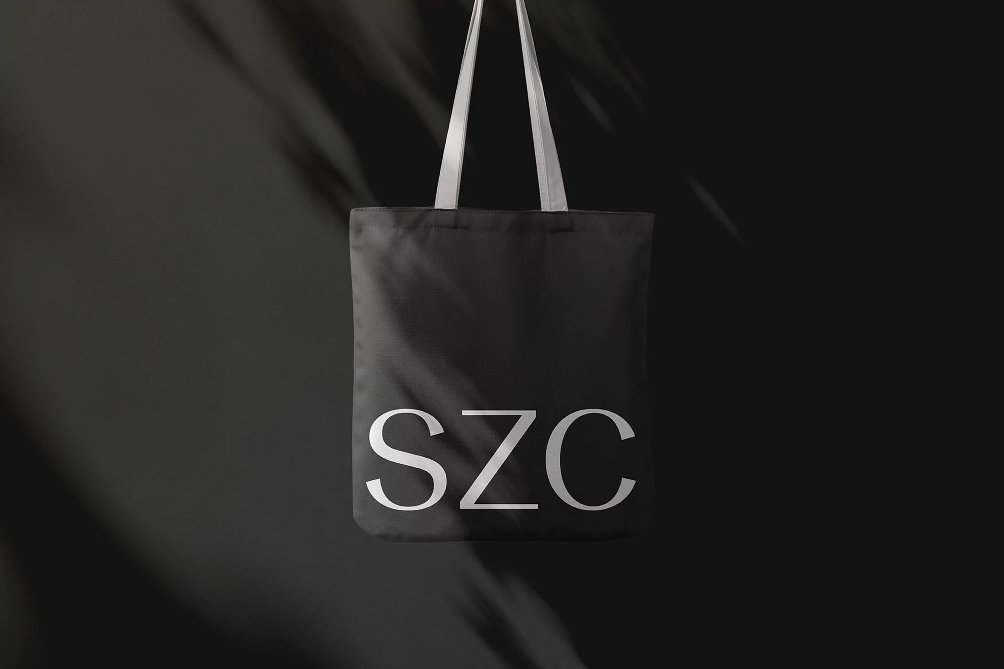 Elegant studio shot of a black tote bag with 'SZC' in white letters, against a dark gray backdrop with soft shadow patterns.