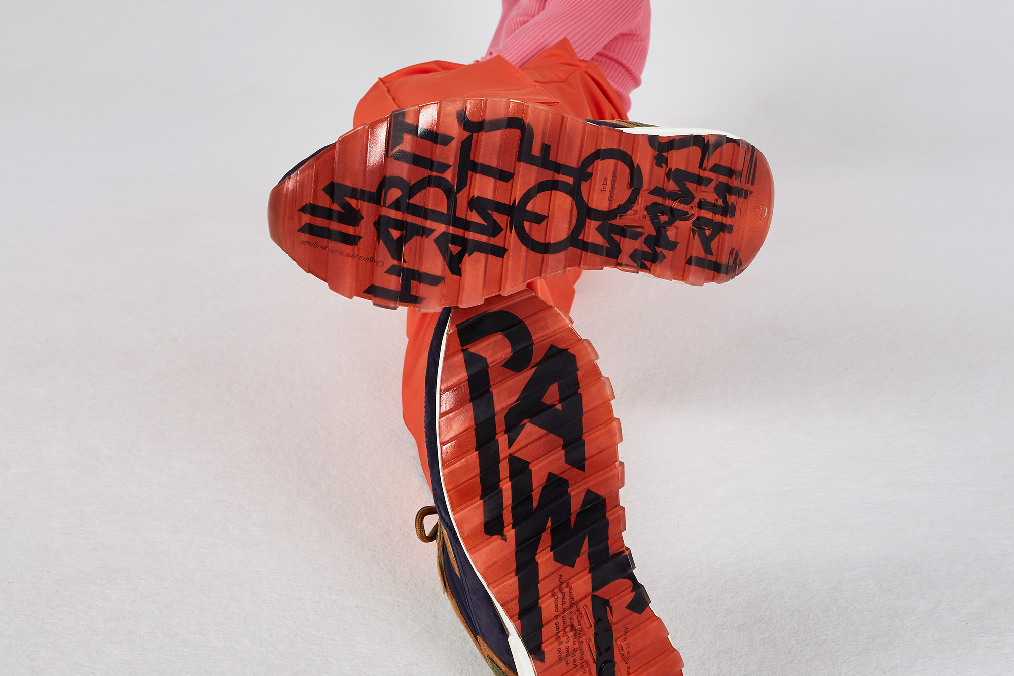 Elevated view of fashion sneakers with coral soles showing brand messaging, orange trousers, and a pink ribbed top on a white background.