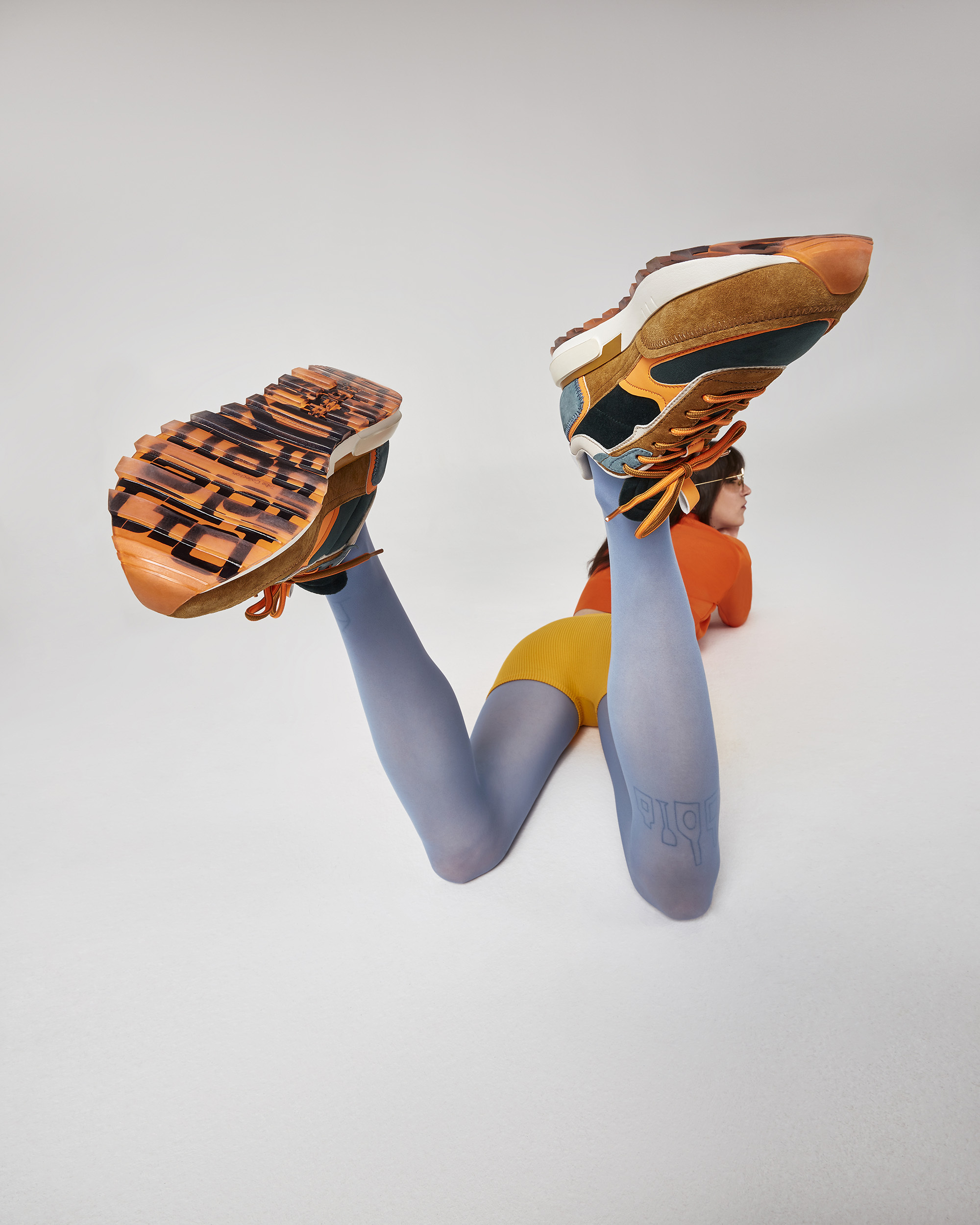 Eye-catching studio shot of orange and brown sneakers held by a person lying on their back, wearing a colorful outfit against a white backdrop.