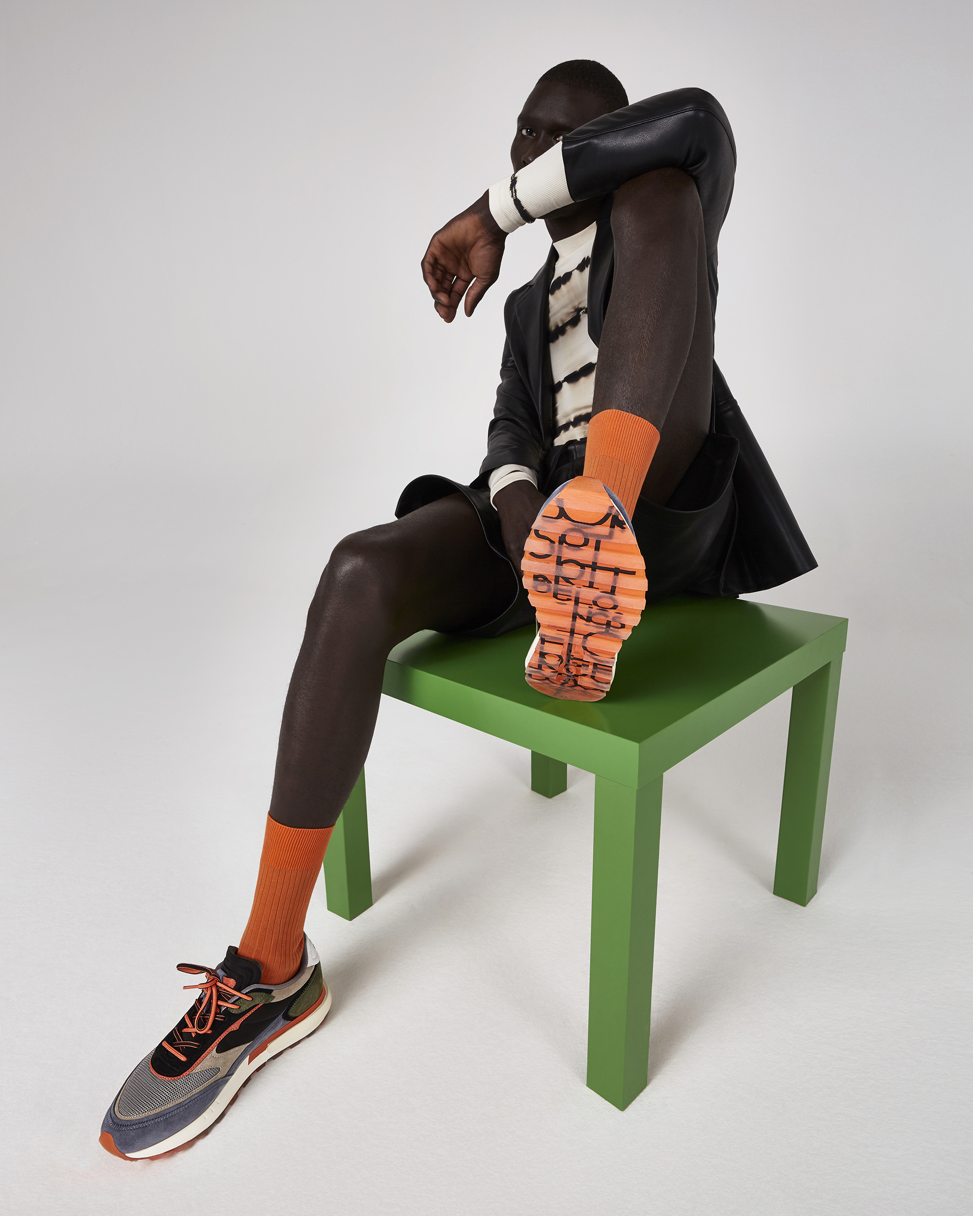 Studio shot of a dark-skinned male model wearing blazer, shorts, and orange socks, sitting on a green table next to a sneaker.