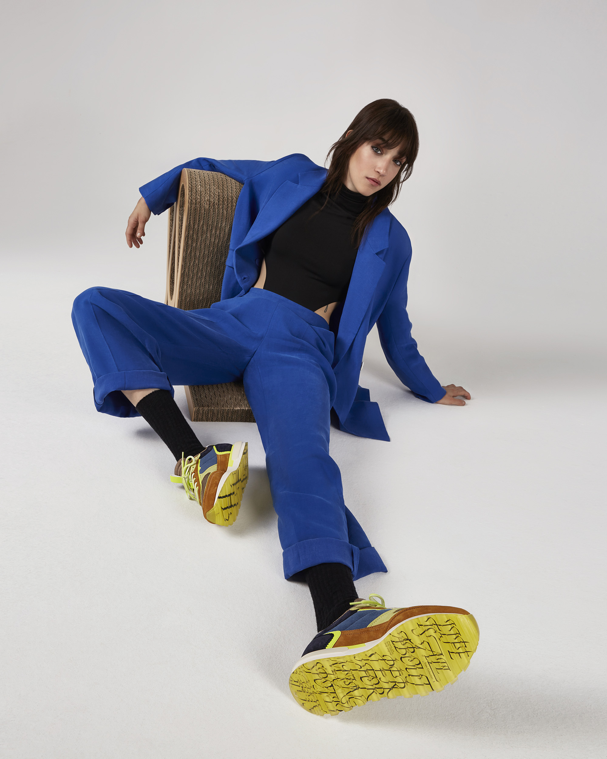 A brunette woman poses in a photo studio, wearing a blue pantsuit, black top and colorful sneakers, sitting on a cardboard chair.