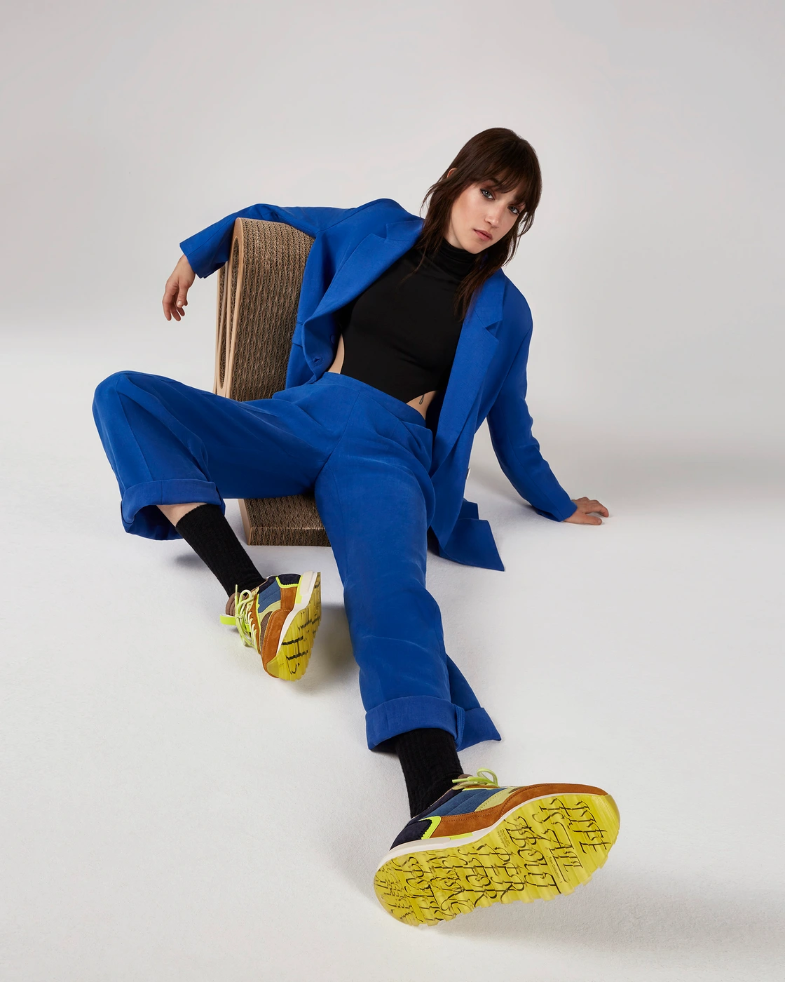 Woman in a cobalt blue suit and black top wearing patterned sneakers seated against a plain white background.