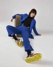 Woman in a cobalt blue suit and black top wearing patterned sneakers seated against a plain white background.