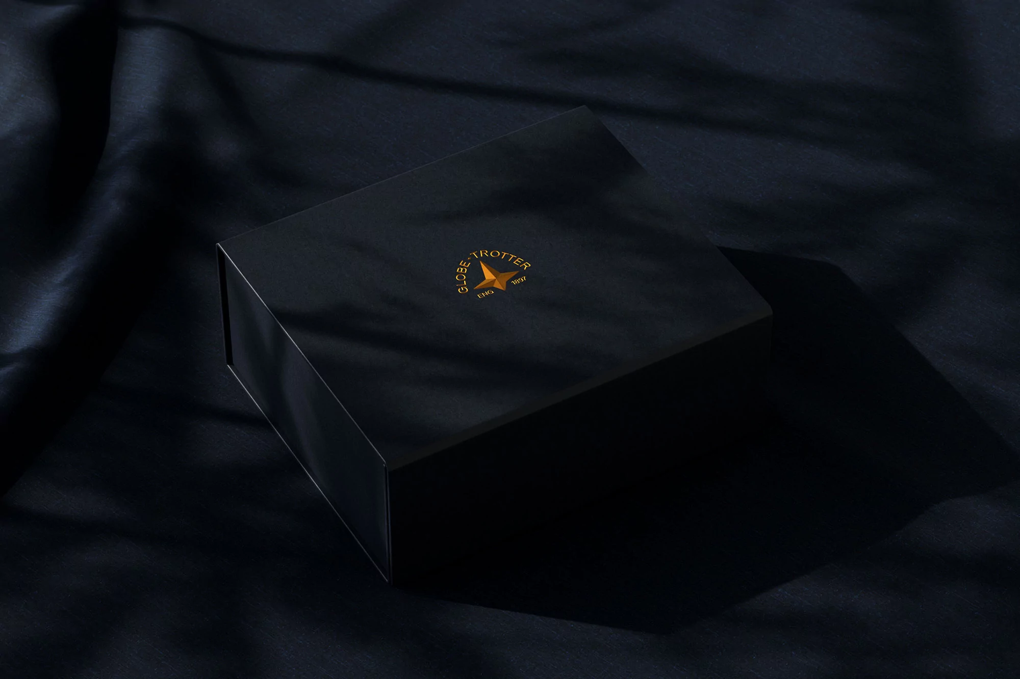 Close-up of a black box with a gold logo on a dark blue satin background with shadow.