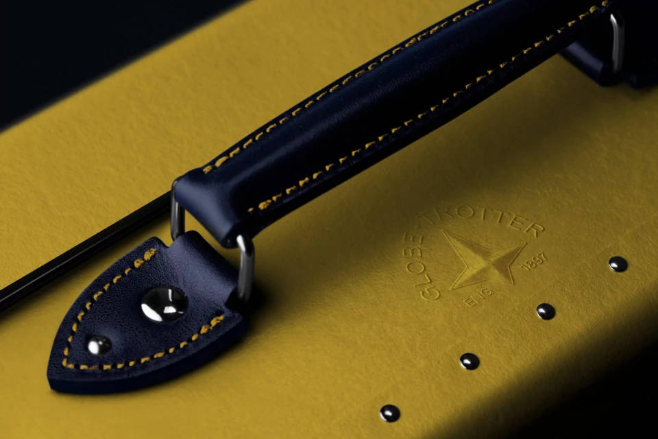 Close-up of a yellow Globetrotter suitcase featuring a navy blue leather handle and silver hardware accents; embossed brand logo visible.