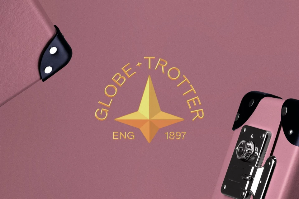 Close-up of a pink vintage Globe-Trotter suitcase featuring black corner protectors and a gold logo with a central star design.