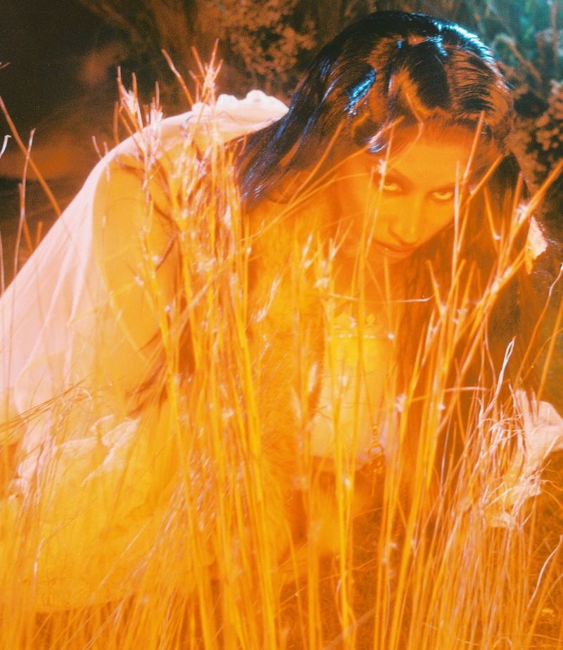 Woman with long hair poses in a white dress surrounded by tall grass, creating a soft, ethereal atmosphere with an orange color grade.