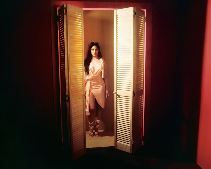 A woman in a light peach dress stands in a doorway with louvered doors. The scene is lit with warm pink tones, creating a soft glow.