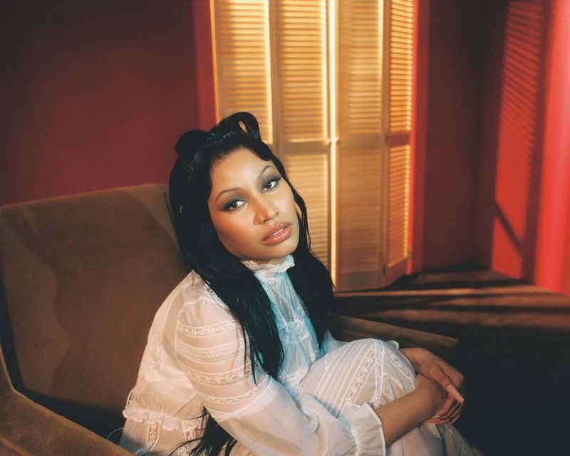 A woman with dark hair poses in a white lace dress, seated on an arm chair in front of a wooden door, captured with a warm color palette.