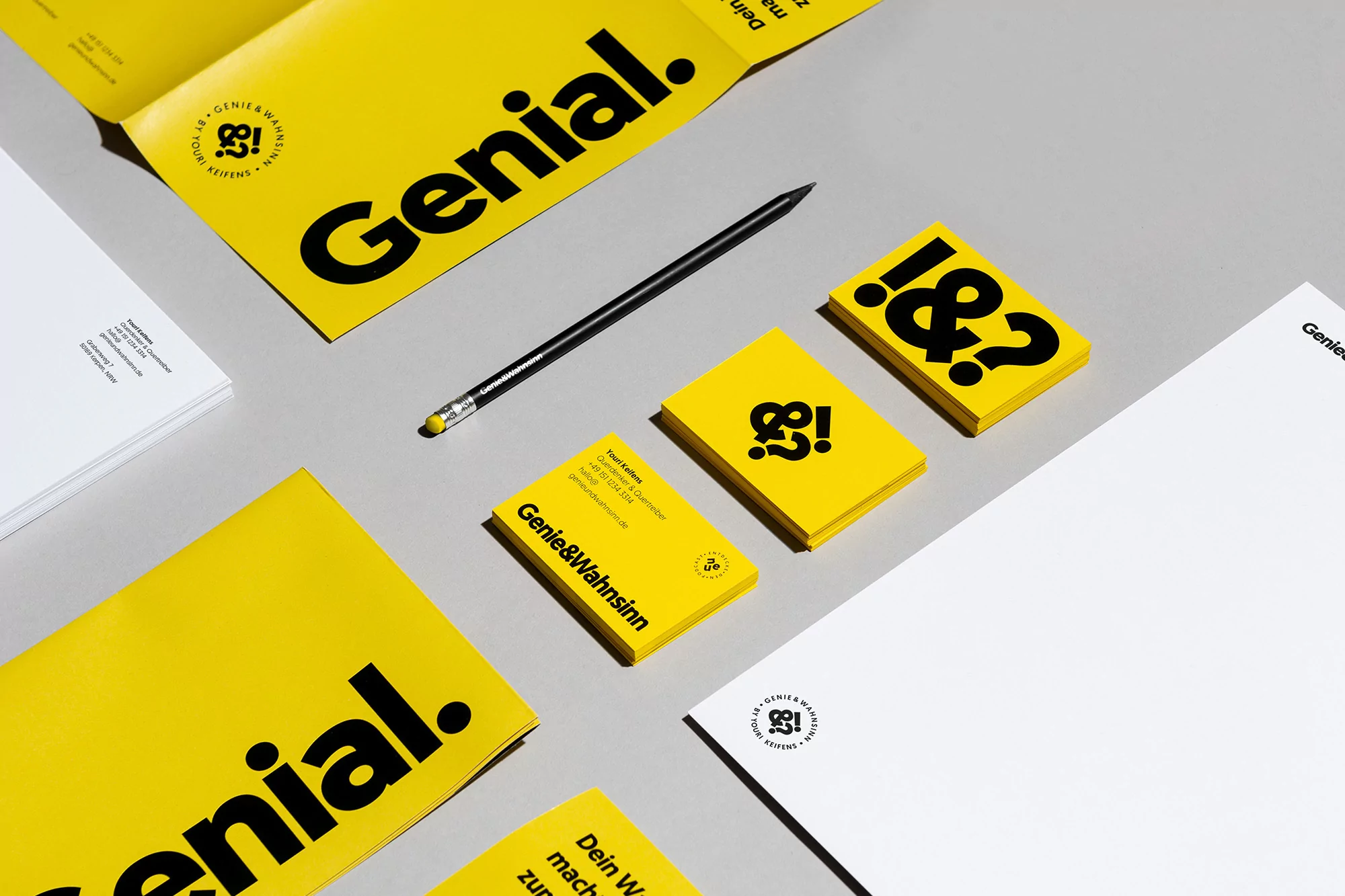 Elevated studio shot of yellow-and-white stationery with brand logos and a black pencil, arranged on a gray surface, for a graphic design concept.
