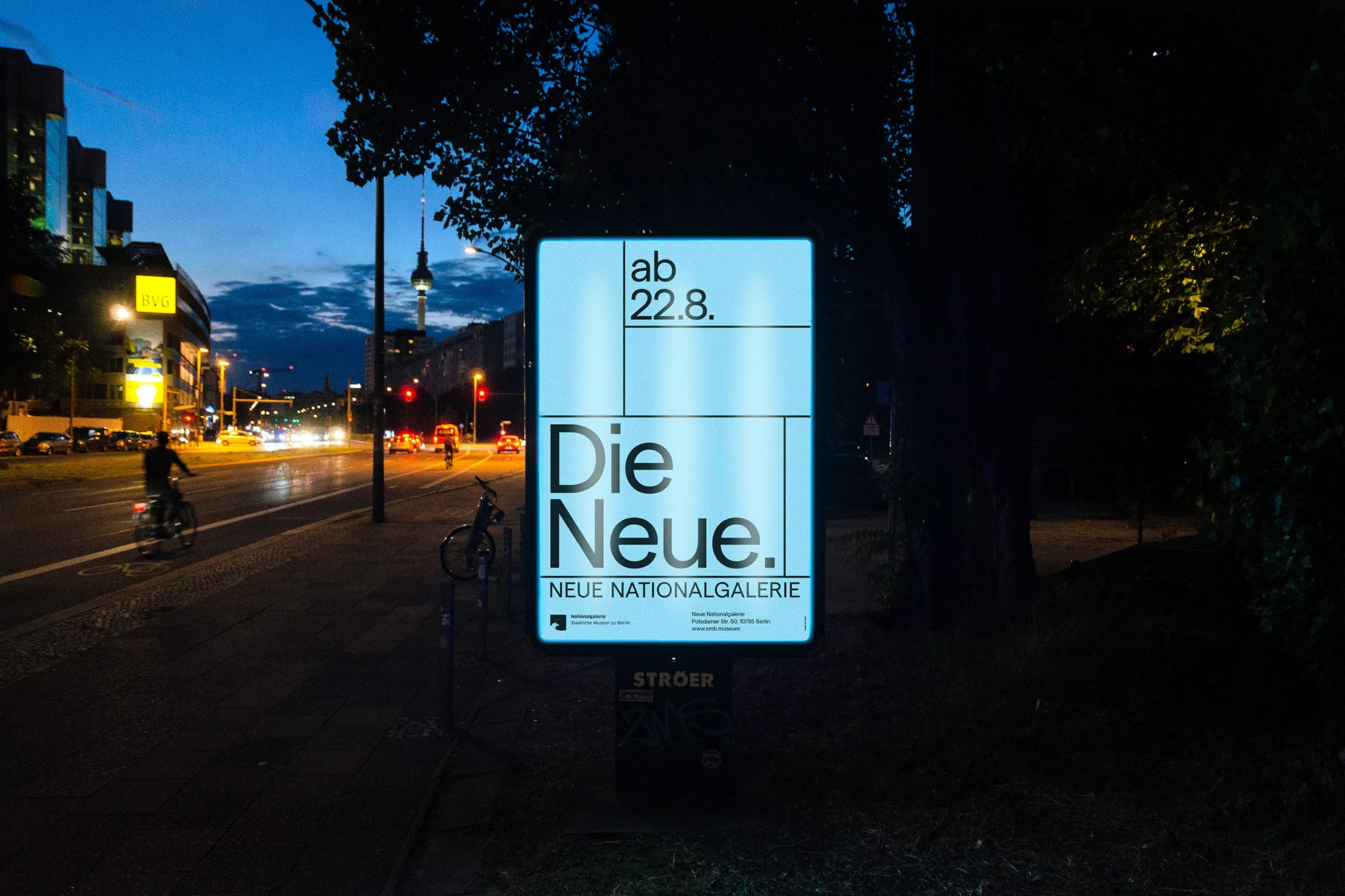Illuminated advertising poster at dusk for Die Neue Nationalgalerie museum with bicyclists and distant Berlin TV Tower.
