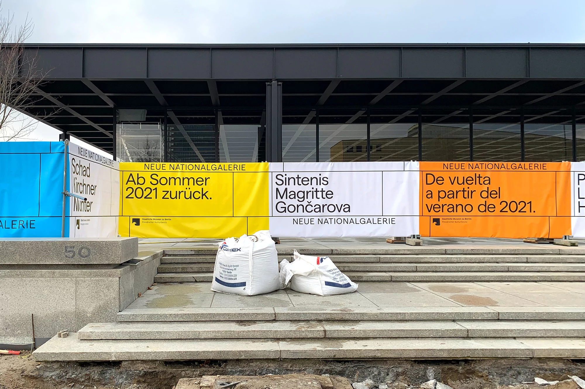 Exterior of Neue Nationalgalerie in Berlin featuring colorful banners announcing upcoming exhibitions, seen during the daytime.