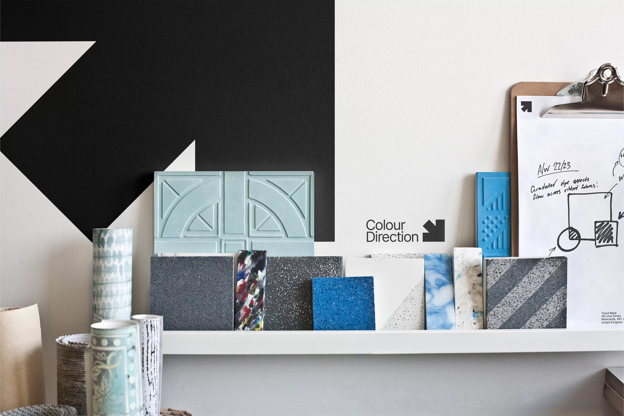 Assorted tiles and design elements arranged on a white shelf against a black and white geometric wall with a clipboard nearby.
